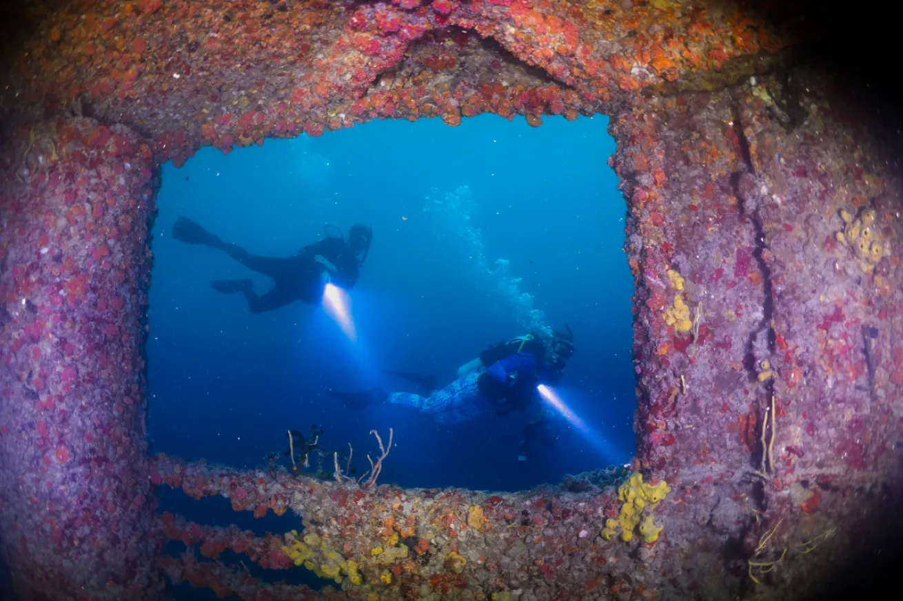 Two divers swimming by a shipwreck with flashlights